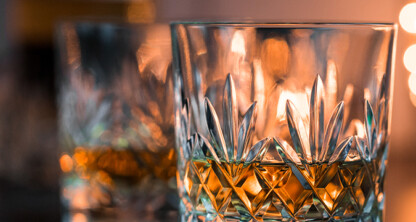 Close-up of a faceted glass filled with amber liquid, likely whiskey, with another glass and blurred warm lights in the background—an inviting scene reminiscent of discussions among Chicago lawyers at a corporate law office.