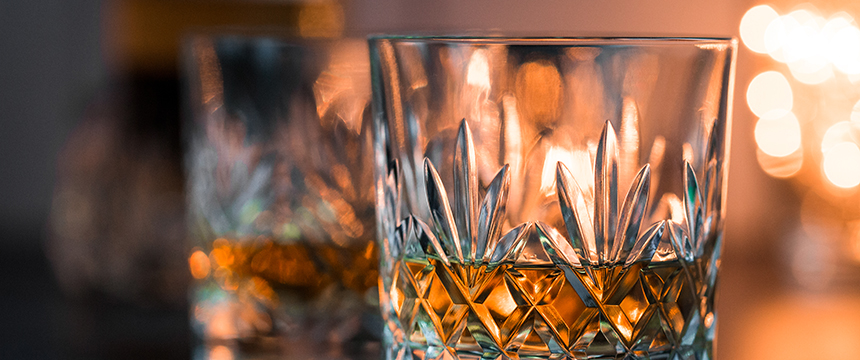 Close-up of a faceted glass filled with amber liquid, likely whiskey, with another glass and blurred warm lights in the background—an inviting scene reminiscent of discussions among Chicago lawyers at a corporate law office.