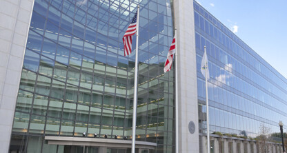 Glass facade of a modern government building resembling leading law offices, with three flagpoles displaying the U.S. flag and two others in front.