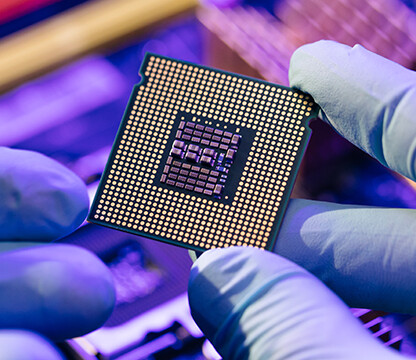 Gloved hands hold a computer processor above a blurred background of electronic components under blue and purple lighting, capturing the advanced technology often utilized by law offices for litigation support.