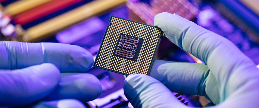 Gloved hands hold a computer processor above a blurred background of electronic components under blue and purple lighting, capturing the advanced technology often utilized by law offices for litigation support.