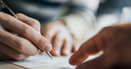Close-up of a person holding a pen and signing a document, with another hand pointing at the paper—capturing a moment often seen in a corporate law office.
