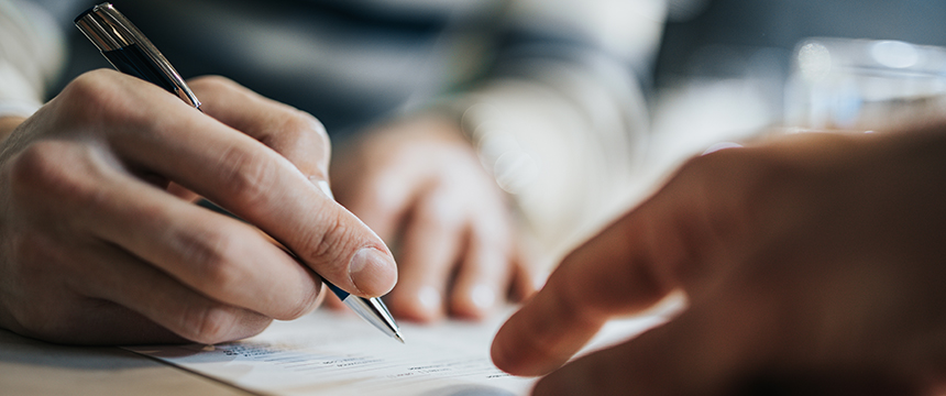 Close-up of a person holding a pen and signing a document, with another hand pointing at the paper—capturing a moment often seen in a corporate law office.