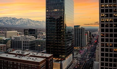 A cityscape at sunset with tall glass buildings, a wide street lined with law offices, and mountains visible in the background under an orange and yellow sky.
