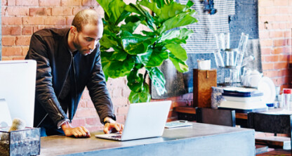 A man stands at a desk in a modern, plant-filled law office, working on a laptop and a desktop computer against an exposed brick wall background, providing litigation support for lawyers in Chicago.