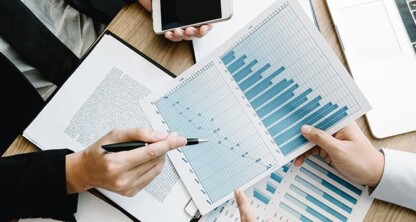 Two lawyers in Chicago review and discuss printed bar and line graphs at a desk with documents, a notebook, and a smartphone, providing litigation support for intellectual property law cases.
