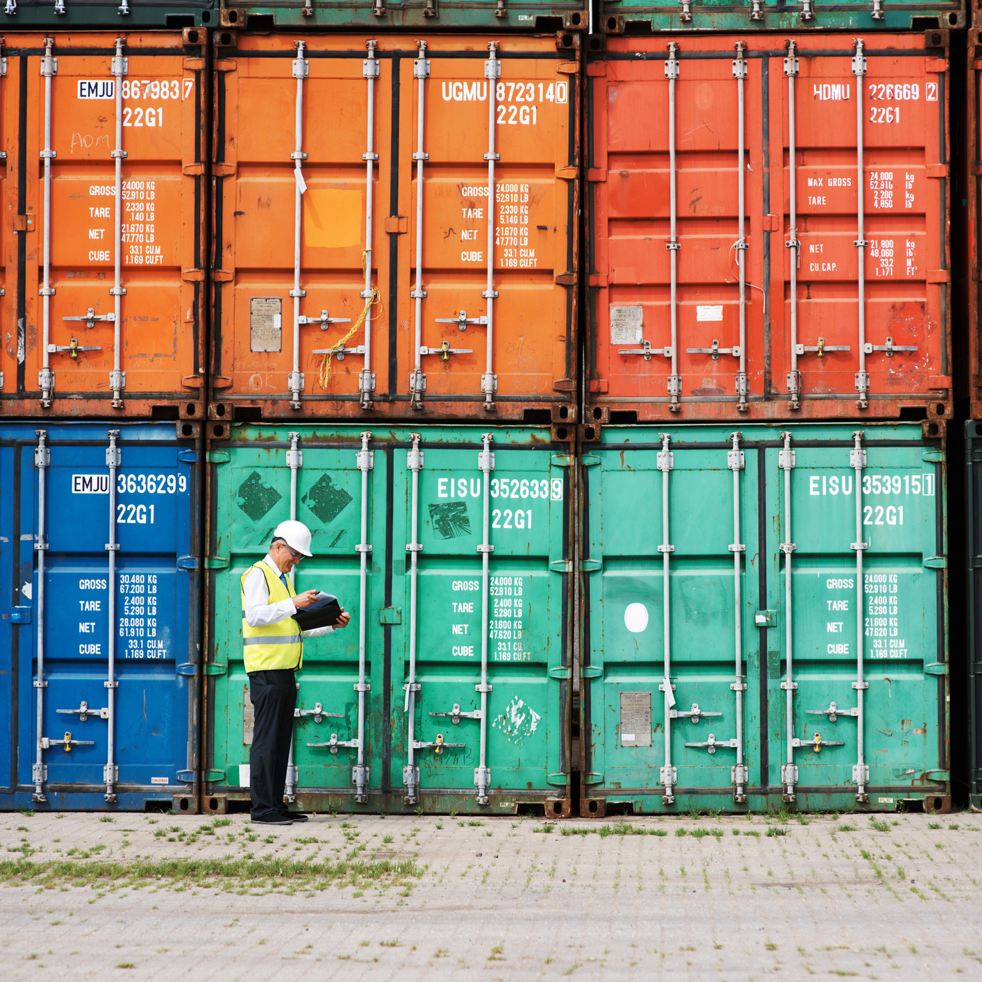 A worker in a safety vest and helmet stands in front of stacked shipping containers, holding a clipboard and inspecting the area near a corporate law office.