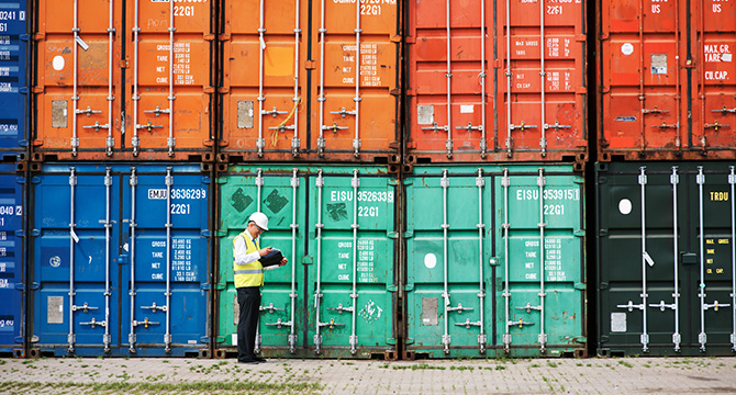 A worker in a safety vest and helmet inspects a clipboard in front of stacked, multicolored shipping containers, ensuring compliance much like Chicago lawyers reviewing critical case documents.