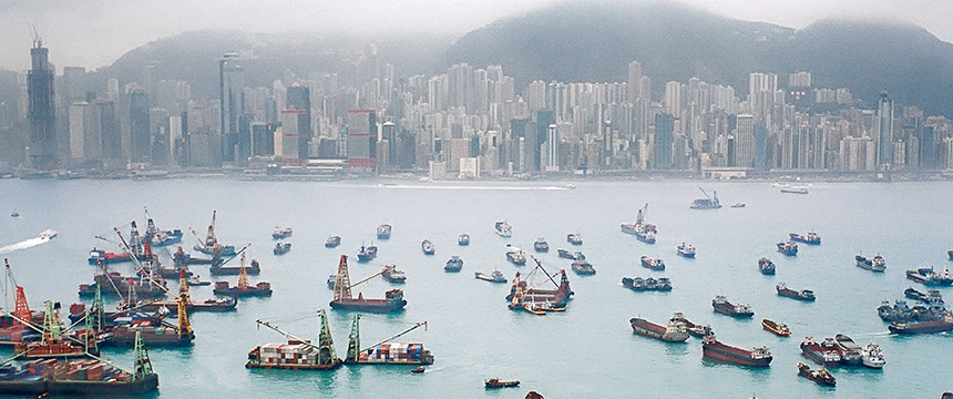 Numerous boats and ships are docked in a busy harbor with a dense city skyline, home to renowned law offices and corporate law office buildings, set against fog-covered hills in the background.