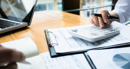 Two people work at a desk in a law office with financial documents, a calculator, a clipboard, a notepad, and an open laptop.