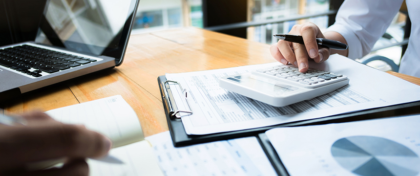 Two people work at a desk in a law office with financial documents, a calculator, a clipboard, a notepad, and an open laptop.