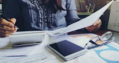 A person sits at a cluttered desk reviewing documents, using a calculator, with a smartphone, eyeglasses, and a calendar nearby—typical of busy law offices specializing in intellectual property law.