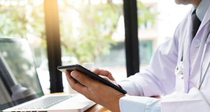 A doctor in a white coat and stethoscope uses a tablet at a desk with a laptop in a well-lit corporate law office.