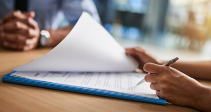 Two people sit at a table while one person, possibly one of the top lawyers in Chicago, reviews and prepares to sign a document on a clipboard with a pen.