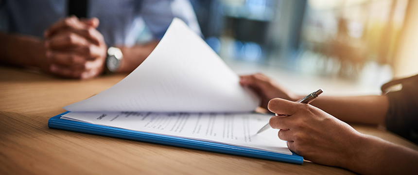 Two people sit at a table while one person, possibly one of the top lawyers in Chicago, reviews and prepares to sign a document on a clipboard with a pen.