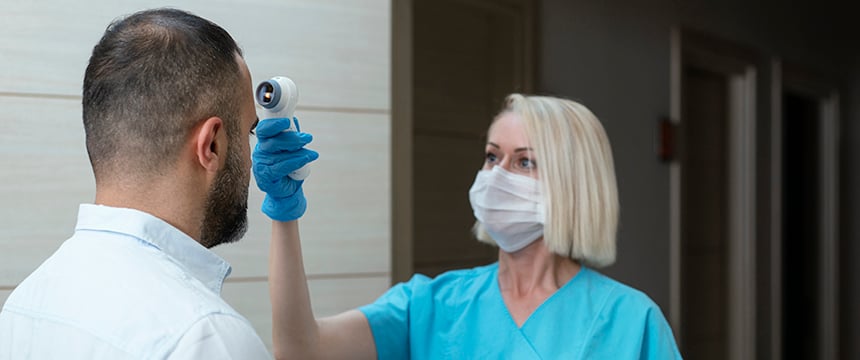 A healthcare worker in scrubs and a mask uses a handheld infrared thermometer to check a man's temperature indoors, as part of safety protocols supported by Chicago lawyers specializing in intellectual property law.