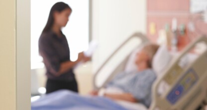 A patient lies in a hospital bed while another person, possibly a chicago lawyer or visitor, stands nearby holding papers. Both figures are out of focus.