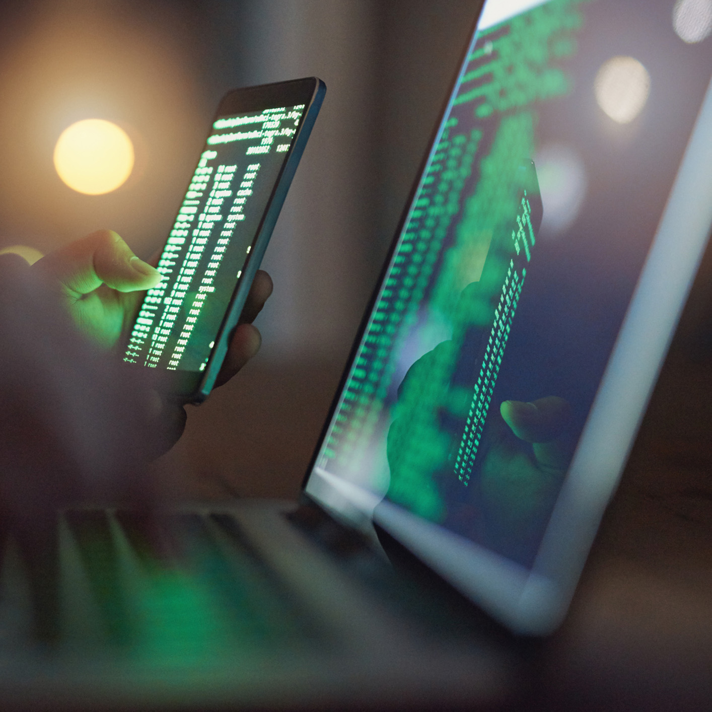 Person holding a smartphone displaying code next to a laptop screen with green code in a dimly lit setting, ideal for showcasing litigation support tools in a corporate law office.
