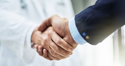 A doctor in a white coat shakes hands with a person in a suit, suggesting a professional agreement or partnership between medical professionals and a corporate law office in a clinical setting.