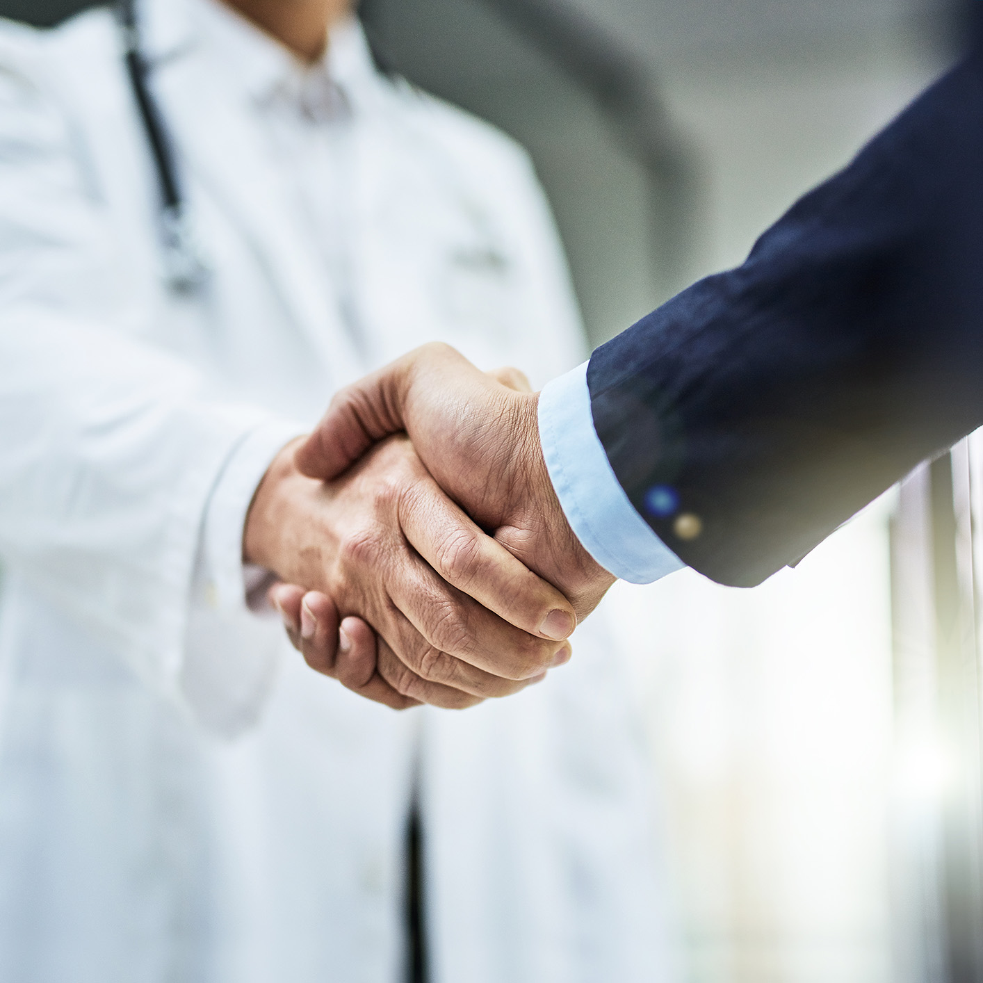 A doctor in a white coat shakes hands with a person in a suit, suggesting a professional agreement or partnership between medical professionals and a corporate law office in a clinical setting.
