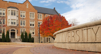 A brick university building with white trim stands behind a stone sign engraved with "UNIVERSITY" and a tree with red autumn foliage, offering an inspiring backdrop often frequented by Chicago lawyers and law offices for litigation support events.