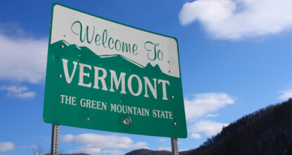 A green and white road sign reads "Welcome to Vermont, The Green Mountain State" against a blue sky with clouds and distant mountains—an inviting view near a leading corporate law office known for its litigation support.