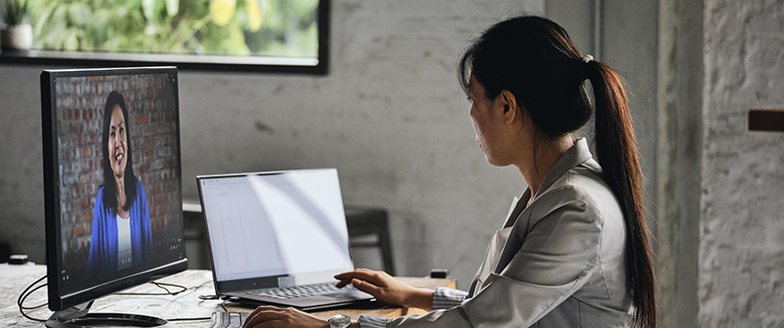 A woman sits at a desk using a laptop, talking to another woman on a video call displayed on a monitor, in a modern corporate law office of Chicago lawyers.
