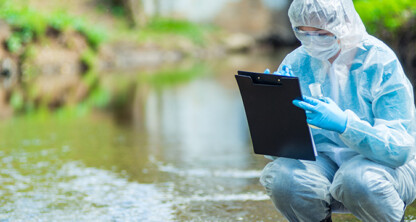 A person in protective gear squats by a river, holding a test tube and writing on a clipboard, perhaps gathering data for lawyers in Chicago investigating environmental compliance cases.