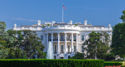 The White House, a large white neoclassical building, stands behind a fountain and neatly trimmed hedges, much like a dignified corporate law office, surrounded by trees under a clear blue sky.