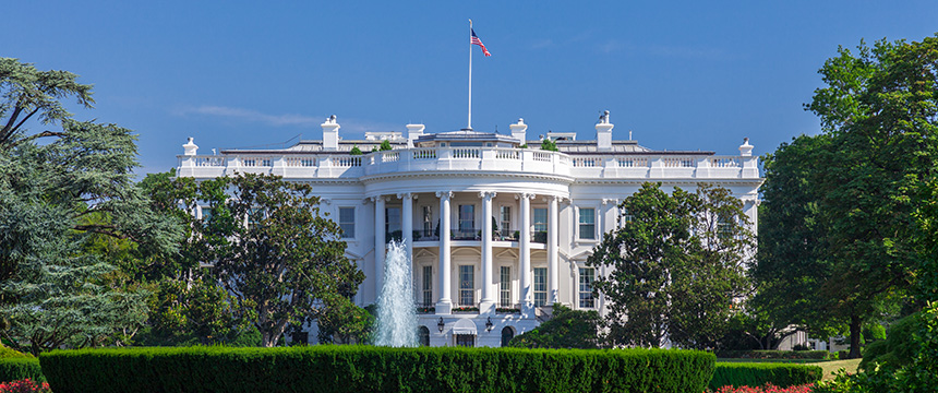 The White House, a large white neoclassical building, stands behind a fountain and neatly trimmed hedges, much like a dignified corporate law office, surrounded by trees under a clear blue sky.