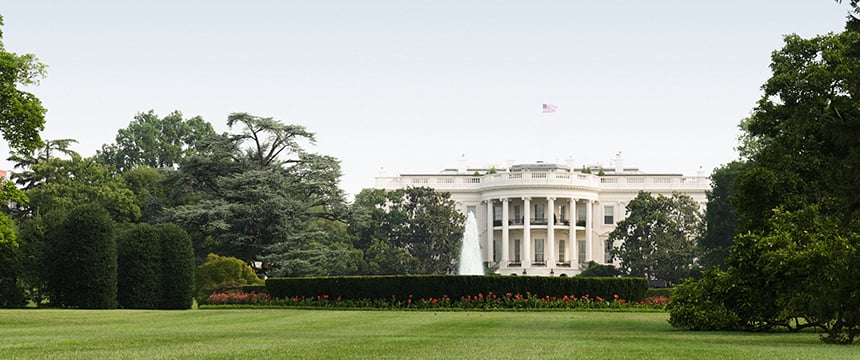 The White House is seen from a distance, surrounded by green lawns, trees, and hedges—a landmark familiar to those in corporate law offices or engaged in intellectual property law, with a fountain and the U.S. flag visible on the roof.