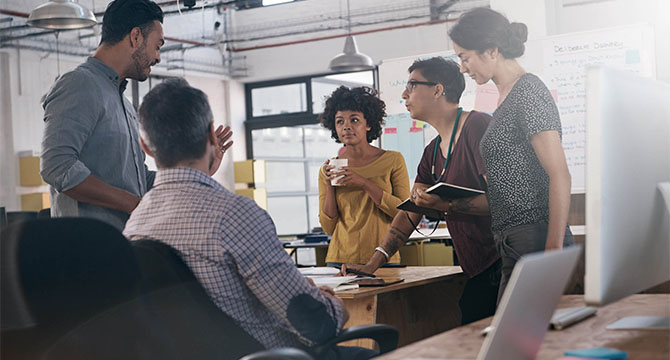 Five people are having a discussion in a modern corporate law office, with one person speaking while others listen and take notes; a whiteboard is visible in the background.