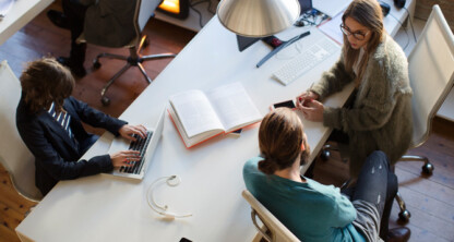 Three people sit around a white desk with computers, a book, and headphones, engaged in work or discussion in a modern law office focused on litigation support.