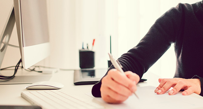 Person writing on paper at a desk in a law office, with a computer, keyboard, and office supplies in the background. Only the person's hand and arm are visible, suggesting work related to intellectual property law or litigation support.