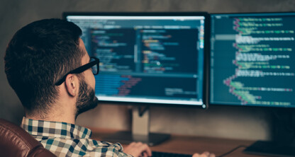 A person wearing glasses sits at a desk in a corporate law office, working on a computer with two monitors displaying lines of code.