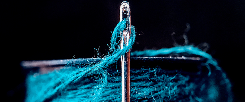 Close-up of a needle with blue thread partially threaded through its eye, with the spool of blue thread in the background—an image symbolizing precision and care, much like litigation support in law offices.