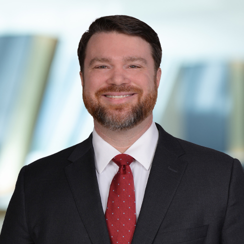 A man in a dark suit, white shirt, and red tie smiles at the camera with a blurred background of vertical shapes, evoking the professional environment of a corporate law office.