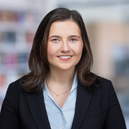 A woman with shoulder-length brown hair, wearing a black blazer and light blue shirt, smiles at the camera in a blurred office setting, reflecting the professionalism of law offices providing litigation support.