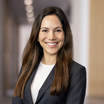 Woman with long brown hair in a dark blazer and white top smiling, standing in a blurred indoor hallway with neutral colors, suggesting a professional environment such as law offices specializing in intellectual property law.