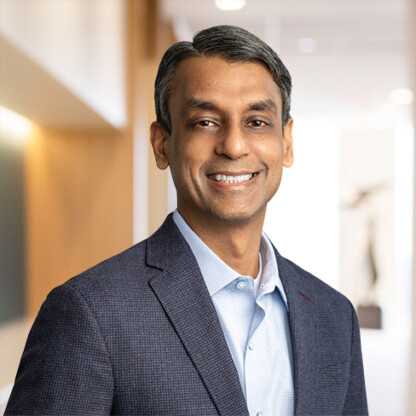 A man in a business suit stands indoors, smiling at the camera, with a blurred corporate law office hallway in the background.