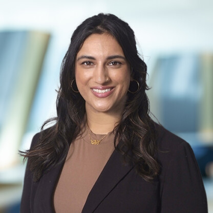 A woman with long dark hair wearing a brown top, black blazer, hoop earrings, and a necklace, smiling at the camera in a blurred corporate law office setting.