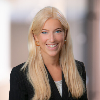 A woman with long blonde hair, wearing a black blazer and white blouse, smiles at the camera against a blurred indoor background in a corporate law office.