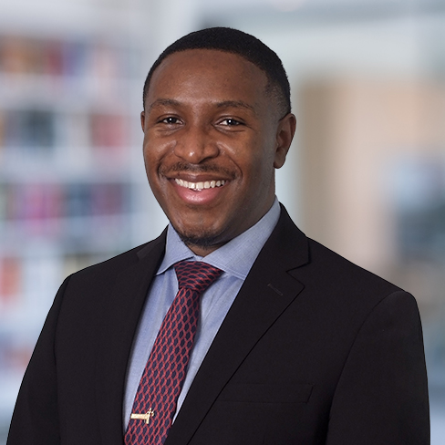 A man in a suit and tie smiles at the camera in an office setting, with blurred shelves typical of law offices in the background.