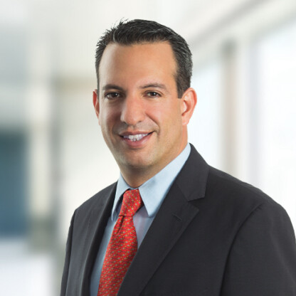 Man in a dark suit with a light blue shirt and red tie, smiling, posed in a bright, blurred office—ideal for representing Chicago lawyers or professionals in law offices specializing in intellectual property law.