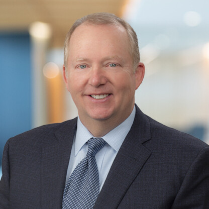 A middle-aged man in a dark suit and patterned tie smiles at the camera in a brightly lit corporate law office.