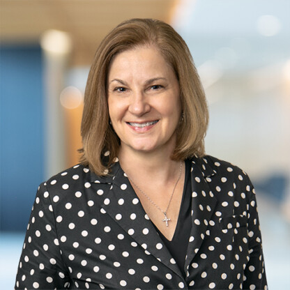 Woman with shoulder-length light brown hair wearing a black blazer with white polka dots, a black top, and a cross necklace, smiling in a blurred corporate law office setting.