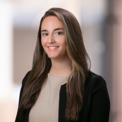 A woman with long brown hair wearing a beige top and black blazer poses for a professional headshot in front of a blurred background, reflecting the polished environment of corporate law offices.