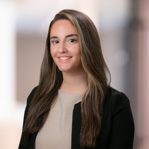 A woman with long brown hair wearing a beige top and black blazer poses for a professional headshot in front of a blurred background, reflecting the polished environment of corporate law offices.
