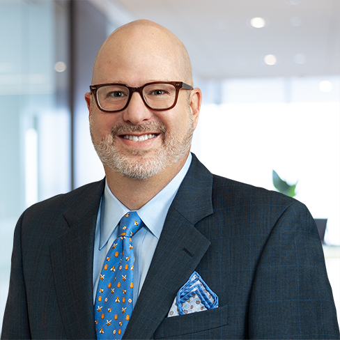 A bald man with glasses, a beard, and mustache, wearing a dark suit, blue shirt, and patterned tie, stands confidently in a modern corporate law office in Chicago.