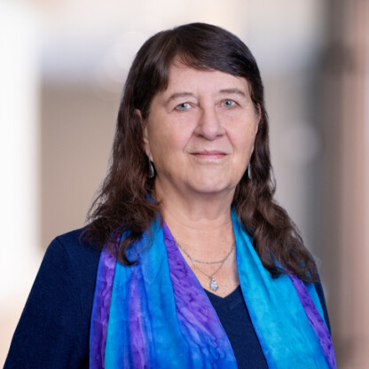 A woman with long brown hair, wearing a blue top, a purple and blue scarf, and a silver necklace, stands in front of a blurred indoor background that could be one of the top law offices for lawyers in Chicago.